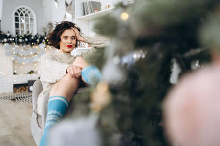 Pretty and happy brunette sitting in warm sweater on the chair with christmas lights behindの写真素材