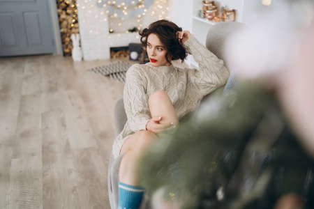 Pretty and happy brunette sitting in warm sweater on the chair with christmas lights behindの写真素材
