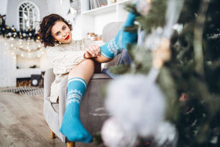 Pretty and happy brunette sitting in warm sweater on the chair with christmas lights behindの写真素材