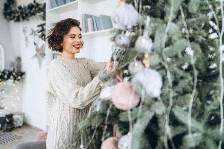 Christmas, New Year. Pretty and happy brunette in warm sweater decorating christmas tree at homeの写真素材