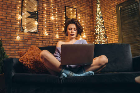 Pretty young brunette at home on the couch using laptop, online education, studying, shopping, working conceptの写真素材
