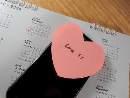 Love you writing on sticker paper with pink heart shape and mobile phone, calendar with marking on February 14thの写真素材