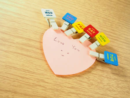 Pink sticky paper, heart shape with happy smiling drawing, love you word, attached colorful days on wooden paper clipsの写真素材