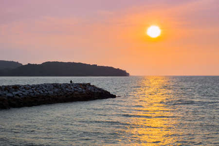 Person sitting on rocks watching sunset on Langkawi island in Malaysia with an orange skyの写真素材