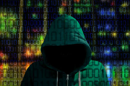 Hacker in a green hoody standing in front of a colored server background with binary streams cybersecurity conceptの写真素材