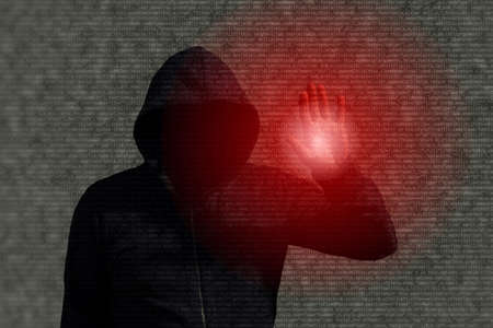 Hooded hacker infiltrating a computer program by touching a screen with red shining light on a computer code backgroundの写真素材