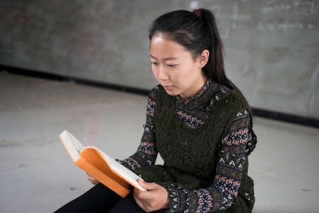 Young student sitting on floor in a classroom and reading a bookの写真素材