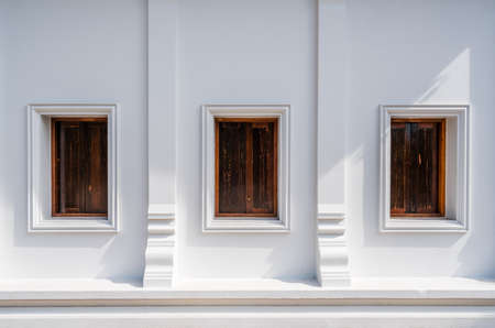 Architecture of Thailand's public temple's wall with three wooden windows, with evening sunlight and shadowの写真素材