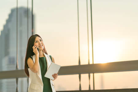 Beautiful young Asian businesswoman use smartphone and digital tablet look upward to copy space, business communication or mobile conference concept, modern office background with sunset and rim lightの写真素材
