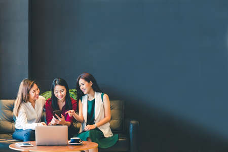 Three beautiful Asian girls using smartphone and laptop, chatting on sofa at cafe with copy space, modern lifestyle with gadget technology or working woman on casual business conceptの写真素材