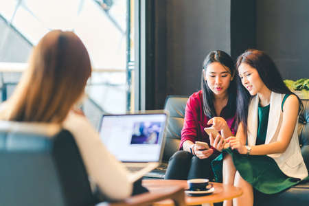 Beautiful Asian girls using smartphone and laptop, chatting on sofa at cafe, modern lifestyle with gadget technology or working woman on casual business conceptの写真素材