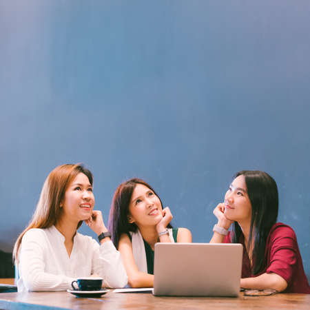 Three beautiful Asian girls looking upward to copy space while working at cafe, modern lifestyle with gadget technology or working woman on casual business conceptの写真素材