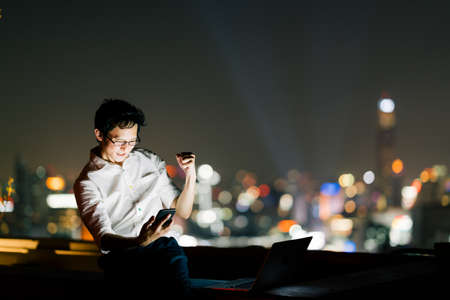 Asian man celebrate with smartphone and laptop computer, success pose. Business, job, or communication gadget technology concept. Night cityscape bokeh background with copy spaceの写真素材
