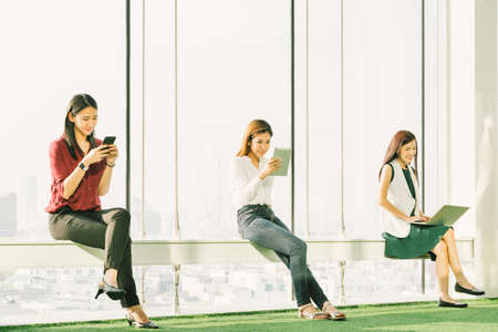 Three Asian girls using smartphone digital tablet and laptop computer in modern office at sunset. Modern lifestyle, information technology, internet gadget, or wireless online communication conceptの写真素材