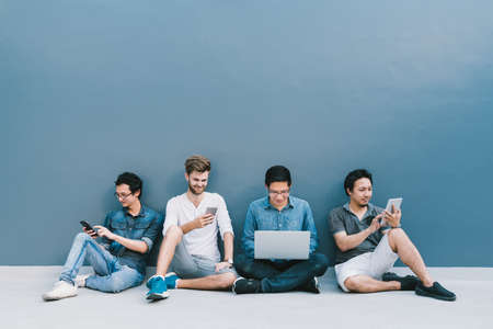 Multiethnic group of four men using smartphone, laptop computer, digital tablet together with copy space on blue wall. Lifestyle with information technology gadget, education, or social network conceptの写真素材