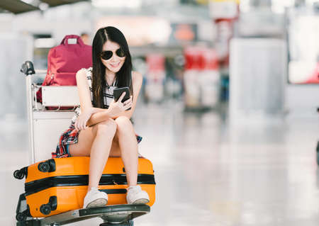 Young Asian girl using smartphone, waiting for flight at airport, sitting on baggage trolley or luggage cart. Mobile communication technology or travel abroad concept, with copy space