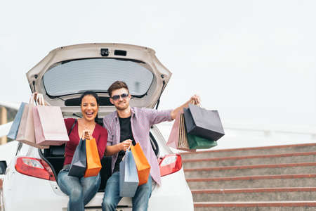 Multiethnic couple with shopping bags, smiling and sitting on white car. Love, casual lifestyle, or shopaholic concept. With copy spaceの写真素材
