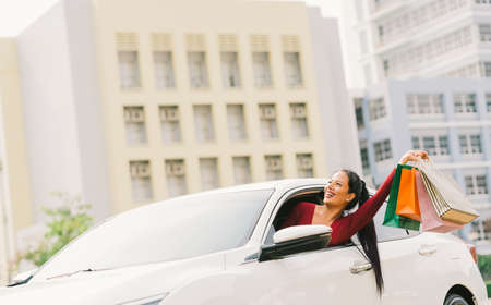 Happy Asian tourist woman raise shopping bags on modern white car, look up at copy space. Shopaholic consumerism activity, retail shop hobby, holiday travel, or casual shopper urban lifestyle conceptの写真素材