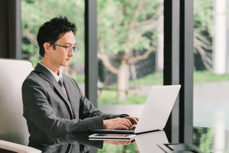 Handsome Asian businessman or entrepreneur at work, using laptop computer and digital tablet in executive office. Leadership, Business communication, startup, or information technology gadget conceptの写真素材