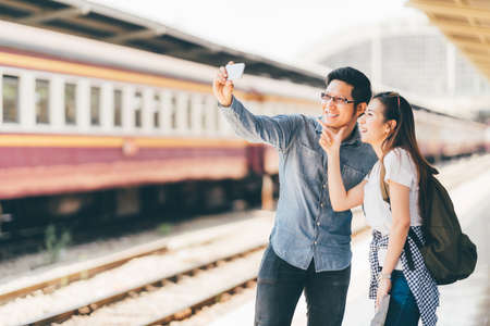 Young Asian couple traveler taking selfie together using smartphone waiting for trip at train station platform in Asia. Backpack travel, Love relationship, holiday vacation or modern lifestyle conceptの写真素材