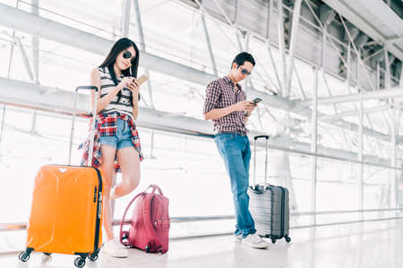 Young Asian man and woman using smartphone checking flight or online check-in at airport together, with luggage. Air travel, summer holiday, or mobile phone application technology conceptの写真素材