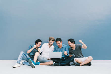 Multiethnic group 4 men celebrate together using laptop computer, sit by blue wall with copy space. College student, information technology gadget, education, social network, or success conceptの写真素材