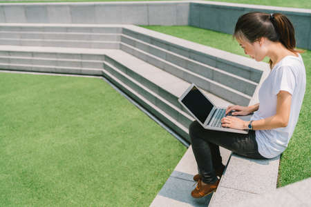 Asian college student or freelance woman using laptop on stairs in university campus or modern park. Information technology, education, or casual business concept. Copy space on green grass yardの写真素材