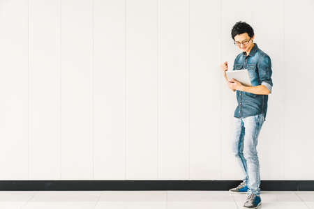 Young Asian man or college student using digital tablet cheering celebrate success, copy space on white wall background. Education, professional job, information technology or mobile lifestyle conceptの写真素材