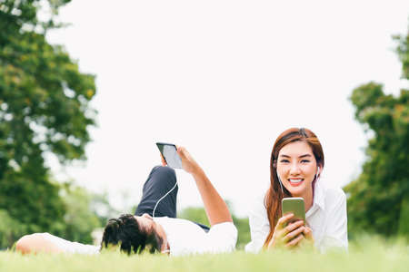 Asian lover couple or college student using mobile phone listen to music together in public park, with copy space. Leisure activity, education, love relationship, marriage, or casual lifestyle conceptの写真素材