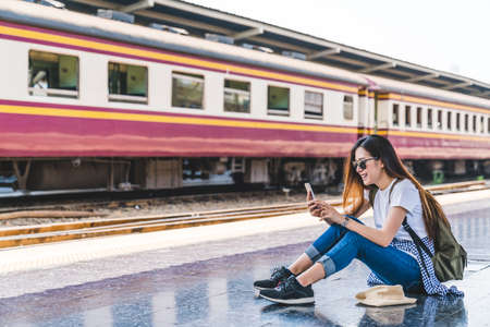 Asian tourist teenage girl at train station using smartphone map, social media check-in, or buy ticket booking. Modern travel app technology, lone traveler, Summer vacation railroad adventure conceptの写真素材