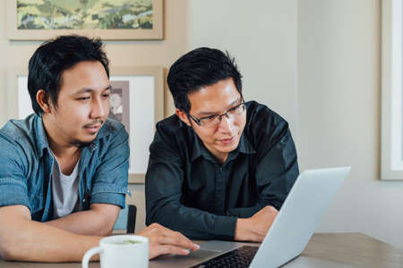 Two Asian businessman or business partner discussing project together using laptop computer at coffee shop. Casual team meeting, teamwork, or startup concept. With copy spaceの写真素材