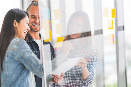Multiethnic diverse group meeting write project plan together on transparent glass board, happy candid smile. Small business startup, teamwork discussion, team building, or strategy brainstorm conceptの写真素材