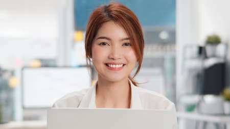 Portrait of young beautiful Asian woman looking at camera and smile, using laptop computer in home office. Happy work life, small business, or internet technology conceptの写真素材