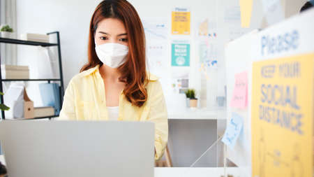 Asian business woman wear face mask, separated from coworker by partition stand. Social distancing at work, new normal office lifestyle after coronavirus lockdown conceptの写真素材