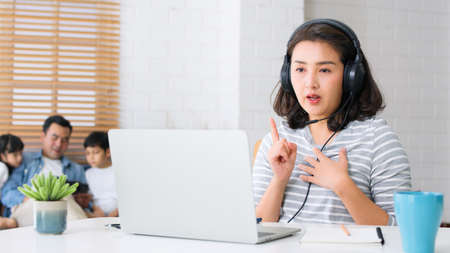 Young Asian mother work at home with family, father, daughter, and son sit together on sofa couch in living room. New normal lifestyle, remote video call online conference meeting conceptの写真素材