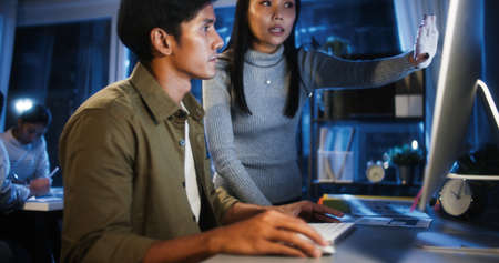 Young Asian business man and woman work late night together on desktop computer with team in home office. Project brainstorm meeting, Small business startup, coworker cooperate teamwork conceptの写真素材