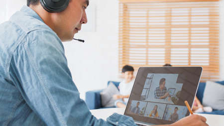 Young Asian father video call conference, online remote meeting with business coworker, with family in living room. New normal lifestyle, social distancing, businessman work at home conceptの写真素材