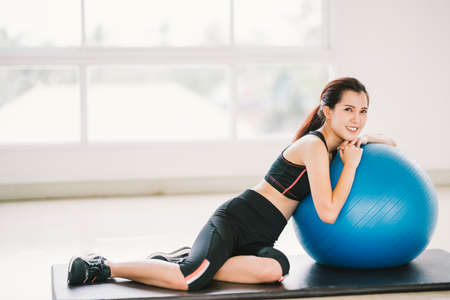 Happy young Asian woman health exercise alone at home gym or sports club, rest on fitness ball. Yoga aerobic class, sport trainer, weight loss, or healthy wellbeing lifestyle concept. With copy spaceの写真素材
