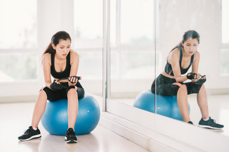 Young, determined Asian woman exercise alone at home gym or sports club, sit on fitness ball with mirror. Yoga aerobic class, sport trainer, weight loss, or healthy wellbeing lifestyle conceptの写真素材