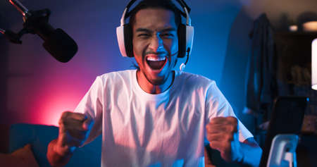 Young Asian man playing online computer video game, colorful lighting broadcast streaming live at home. Ecstatic celebration winning a match. Gamer lifestyle, E-Sport online gaming technology conceptの写真素材