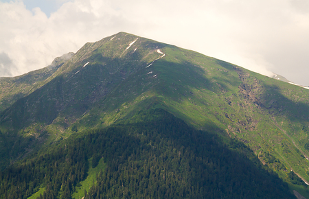 view of the slope of the desolate mountain with steep slopes covered by forest close to the white dense cloudsの写真素材
