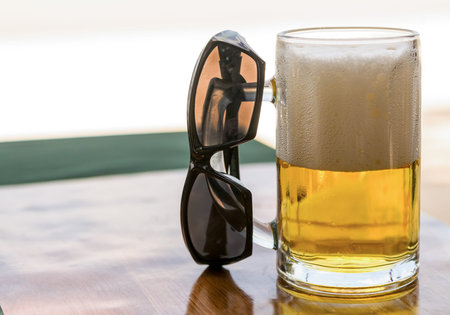 glass mug of golden beer with white high foam and sunglasses on a wooden table