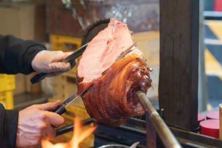 hand with a knife cuts a portion from a knuckle on a skewer, a leg of pork cooked over an open fireの写真素材