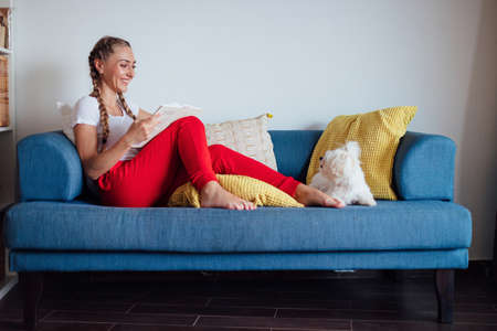 Side view composition of smiling young woman sitting on the couch with her dog and reading book while enjoying weekend at homeの写真素材