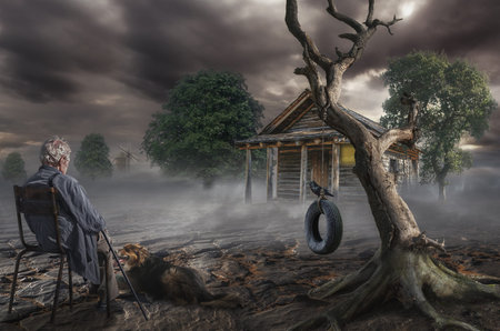 an elderly gray-haired man sits near a dry tree on the spot where was the house of his childhoodの写真素材