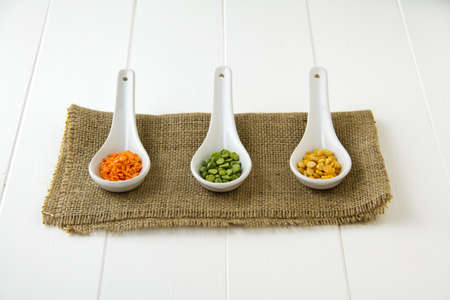 Raw, dried red lentils, green lentils and yellow lentils in three white ceramic spoons, sitting on burlap and white wooden boardsの写真素材