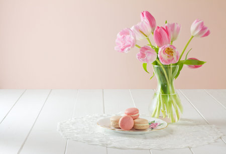 A heap of French macarons (pronounced macaroon, a popular buttercream filled meringue type cookie or biscuit) on a dainty porcelain, rose patterned and gilded plate, on white wood table with lace placemat. Soft pink and white tulips sit to one side. A freの写真素材