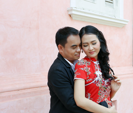 Asian bridegroom hold his bride in red cheongsam dress against pink building backgroundの写真素材