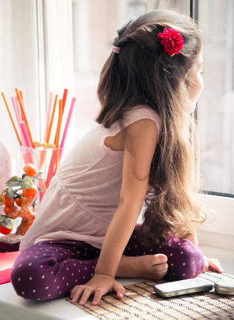 Long-haired curly little girl sitting on window-sill looking out the window.の写真素材