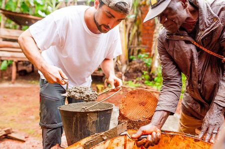 Bangoua, Cameroon - 08 august 2018: young european man in african village doing manual work building water well with concrete in copperation with local old manのeditorial素材